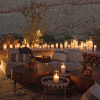 A cozy outdoor evening setup with plush seating wooden tables adorned with flowers and numerous lit candles creating a warm ambiance against a rugged desert cliff backdrop This serene scene is ideal for a romantic or relaxing night under the stars blending nature with elegant decor for a memorable desert experience My local utah