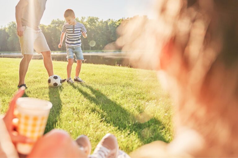 Boy in striped shirt and an adult kick a soccer ball on a sunny park lawn, with blurred spectators in the foreground.