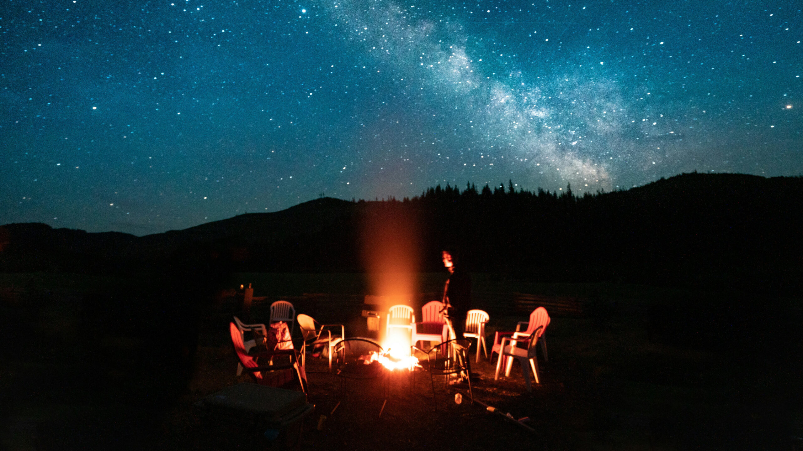 Campfire circle with colorful chairs around a fire under a starry night sky and Milky Way over dark hills.