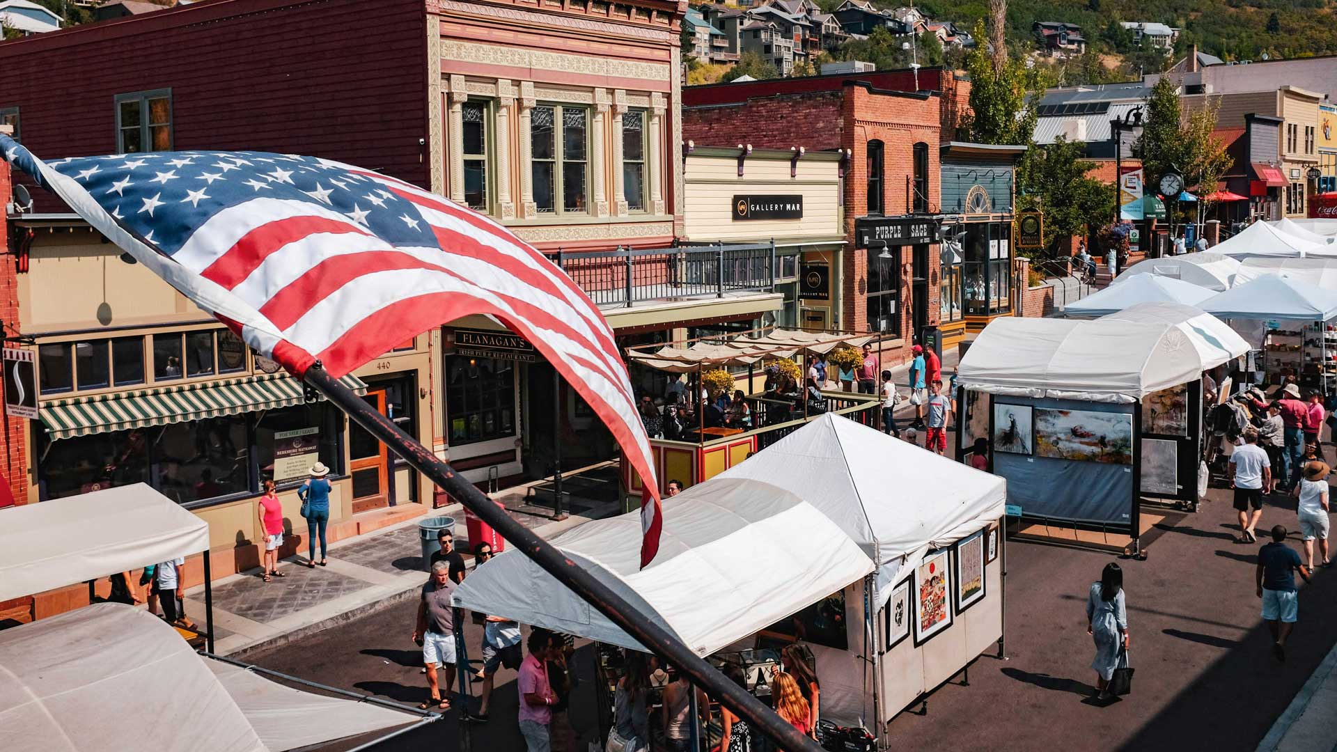 Outdoor street market with white tents, storefronts, and a waving American flag in the foreground.