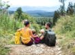 Family of four sits on a rocky forest trail with backpacks, overlooking a green valley and distant mountains.
