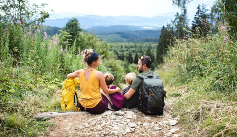 Family of four sits on a rocky forest trail with backpacks, overlooking a green valley and distant mountains.