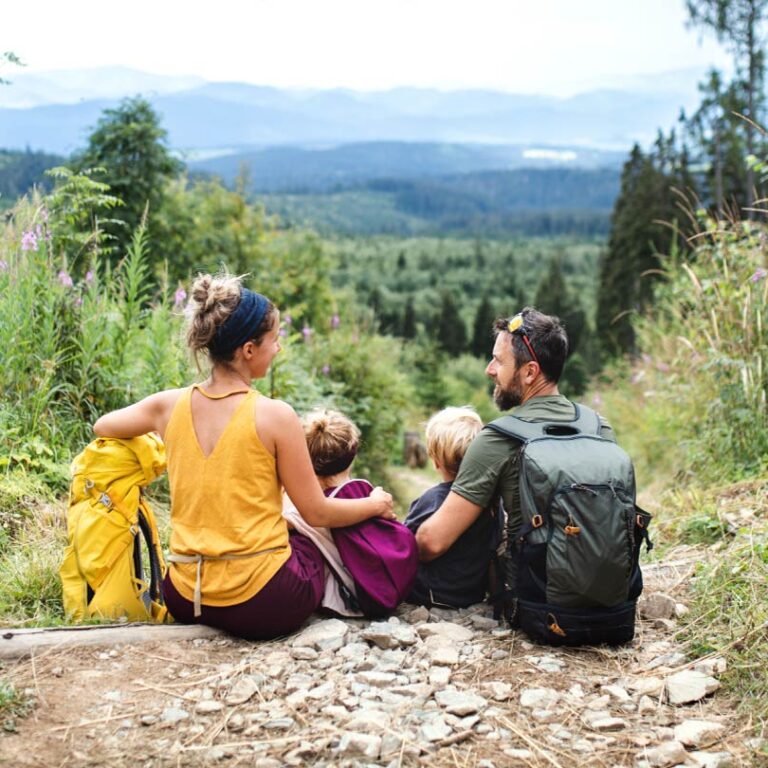 Family of four sits on a rocky forest trail with backpacks, overlooking a green valley and distant mountains.