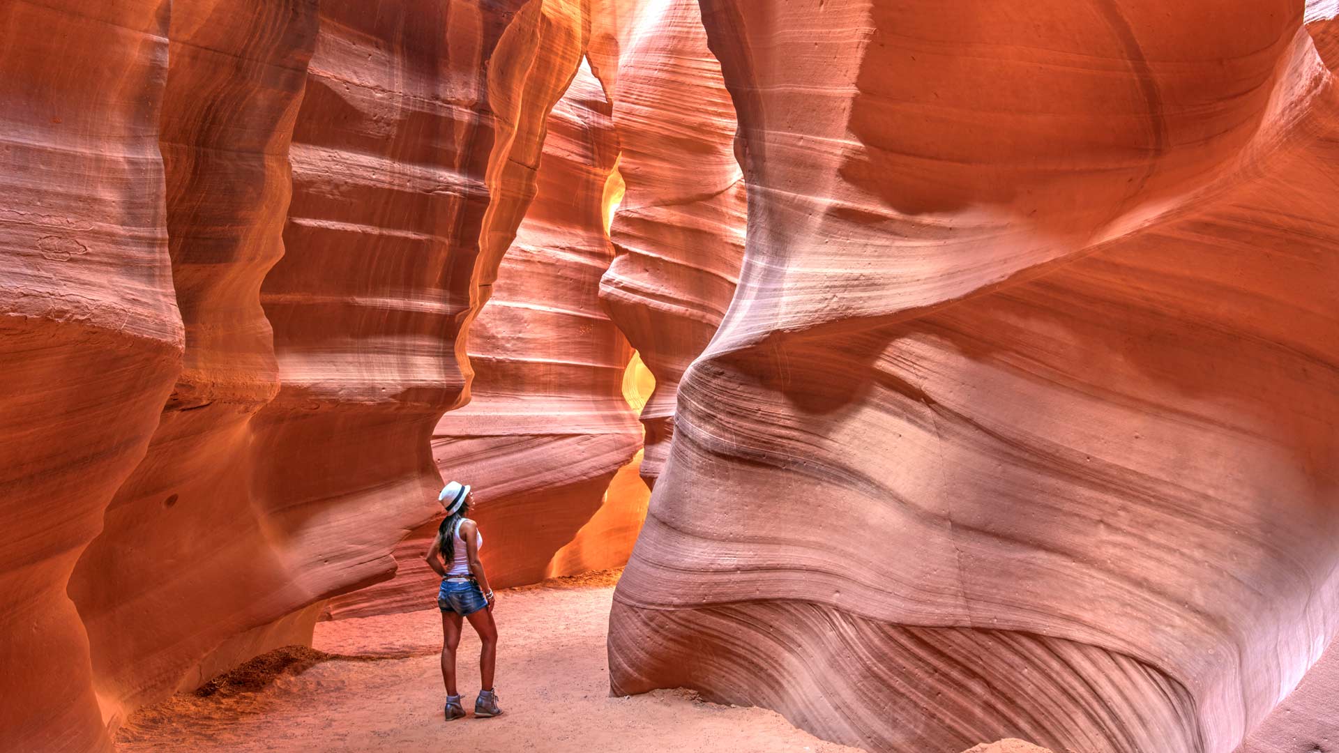 Person wearing a hat exploring a narrow red sandstone slot canyon in warm light.