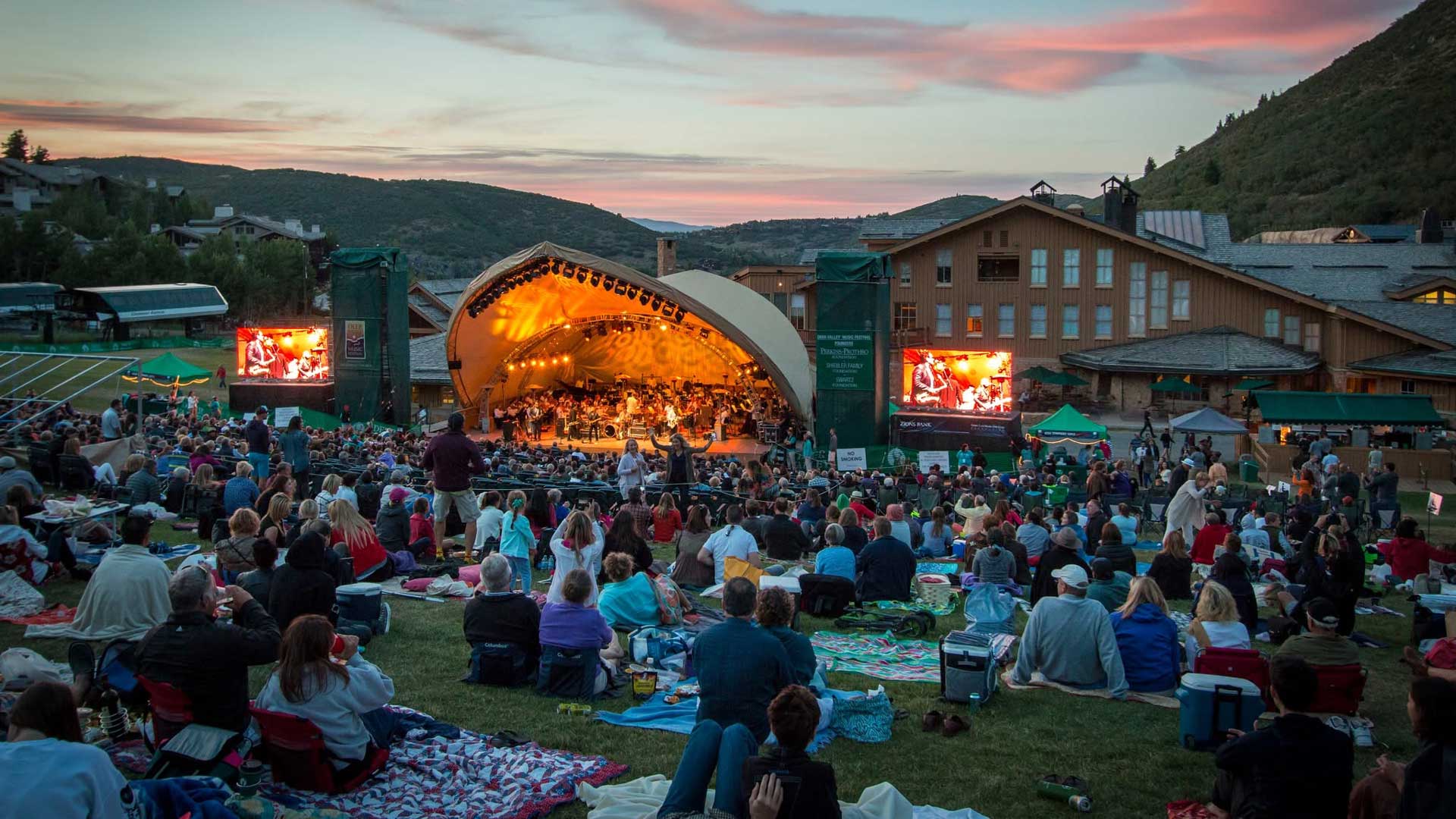 Open-air concert at sunset with a large crowd seated on blankets watching a band perform on a curved, illuminated stage.