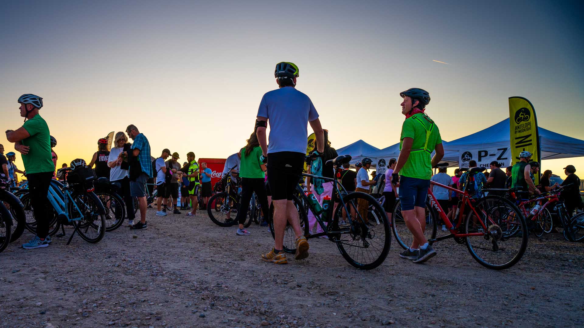 Group of cyclists and spectators gather near tents at sunset during a biking event; bikes lined up and people chat.