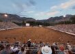 Outdoor rodeo arena plein with dirt track, grandstands full of spectators, and mountain range in the background at dusk.