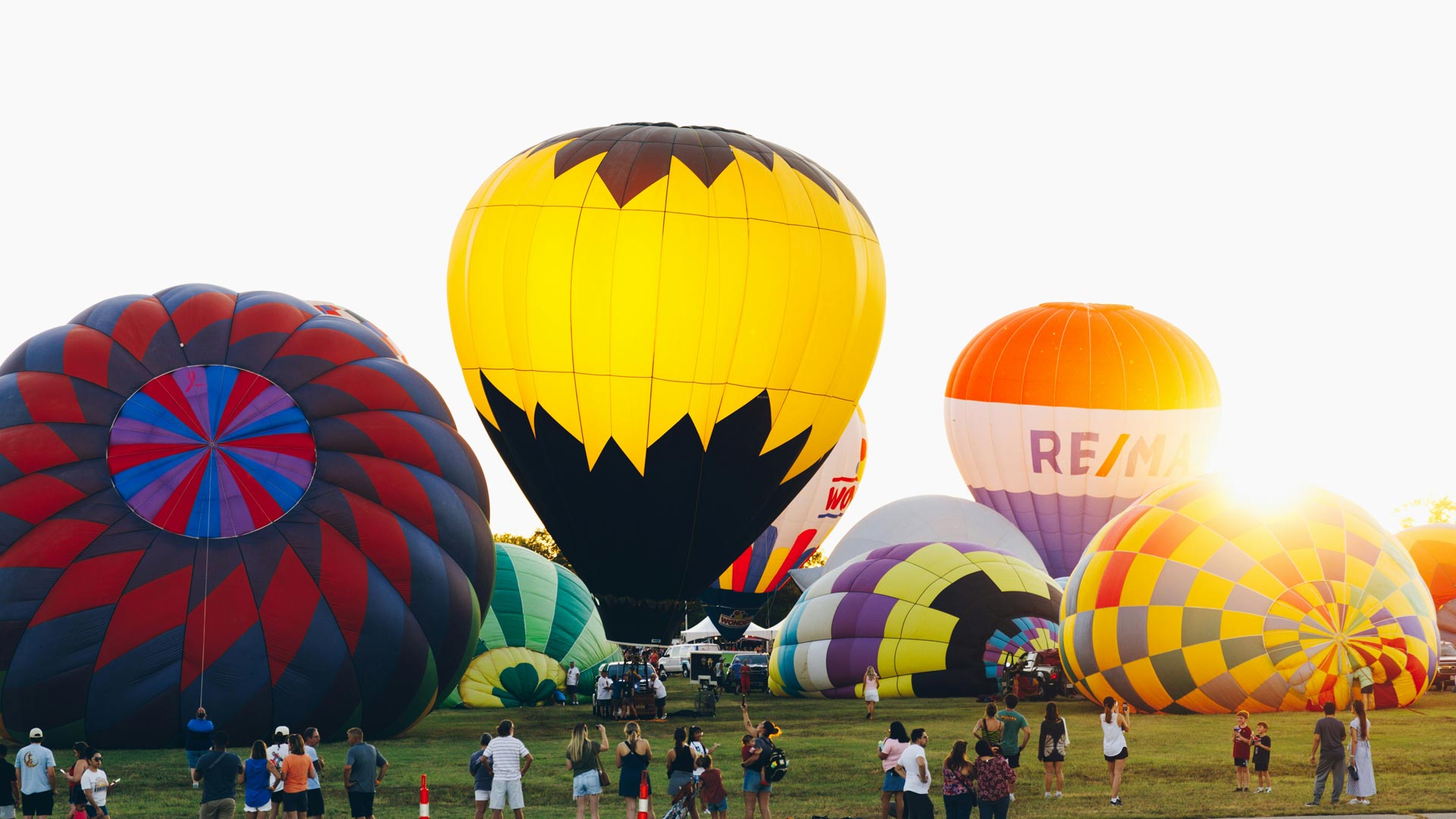 Colorful hot air balloons inflated on a grassy field at a festival, with spectators gathered around.