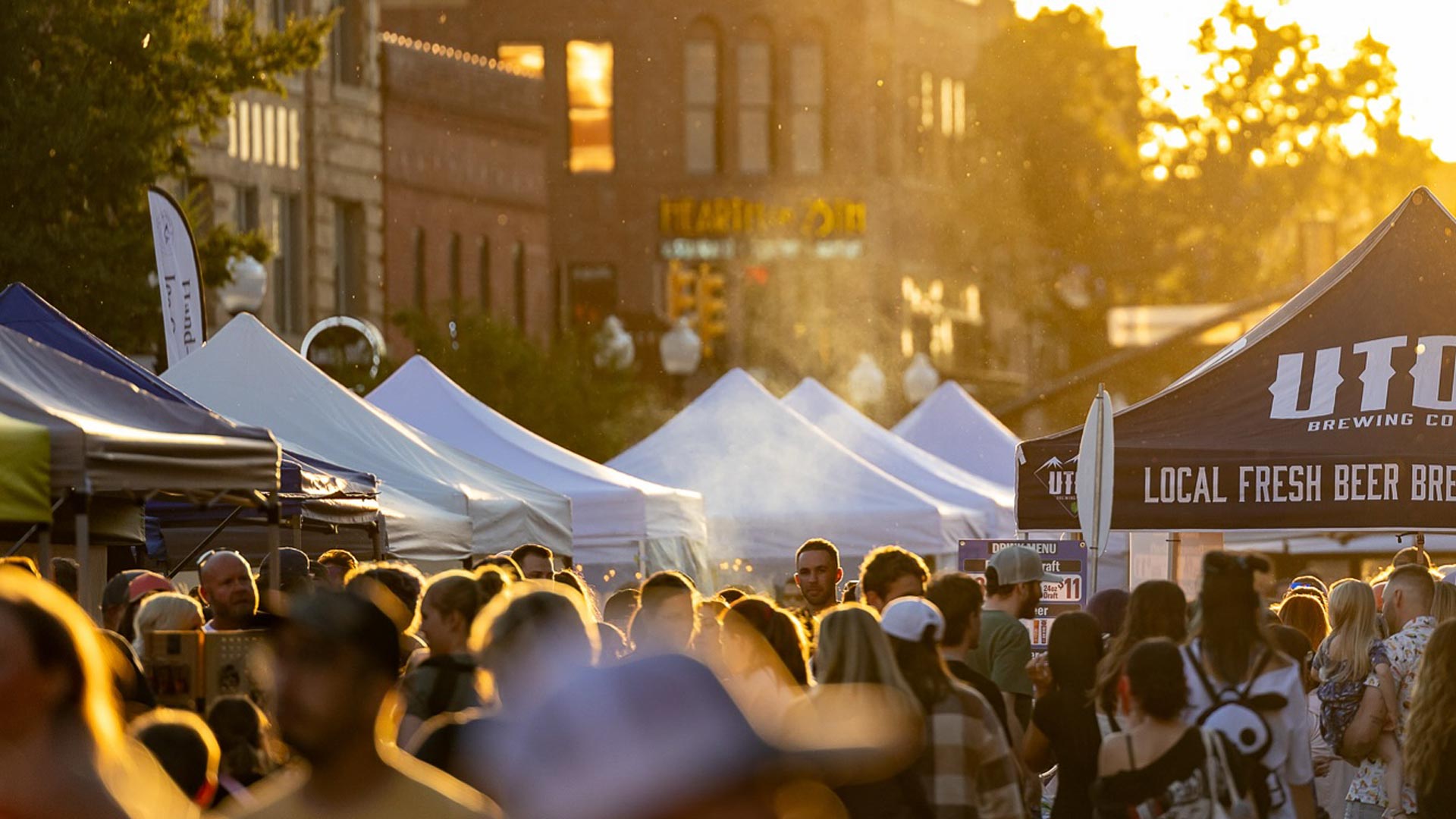 Crowd at an outdoor street festival with white tents and warm sunset lighting.