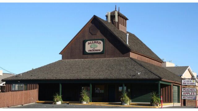 Barn-style Allred Orchards storefront with a vertical fruit-sign (peaches, apples, nectarines) against a clear blue sky.