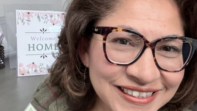 Smiling woman with tortoiseshell glasses and a plaid coat, pink shirt visible; background shows a decorative sign reading 'Welcome'.
