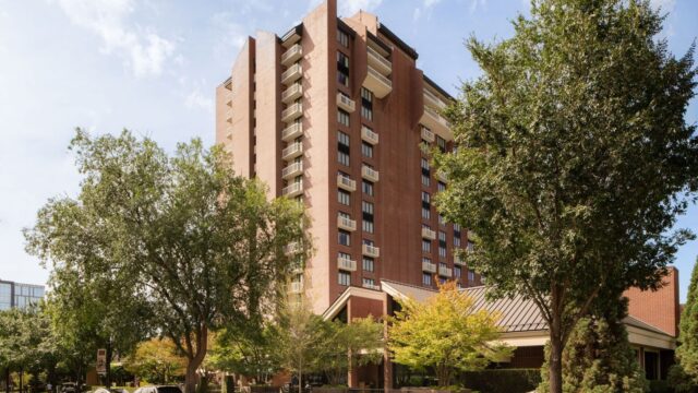 Brick apartment building with balconies, surrounded by trees and a manicured lawn under a blue sky.