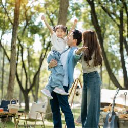 Father lifts daughter while mother watches in a sunny forest campsite; family enjoying outdoor picnic setup nearby.